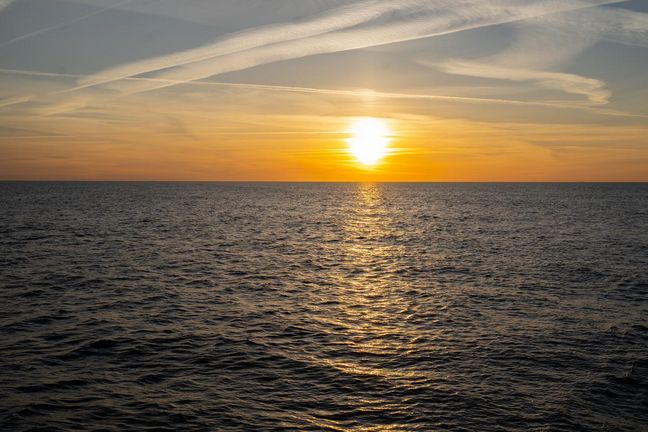 Sonne steht hinter einem gelben Himmel &uuml;ber dem Horizont. Im Vordergrund ist das Meer zu sehen.