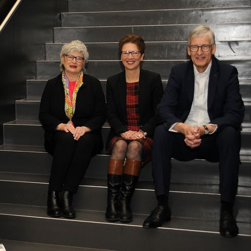 Gruppenbild mit zwei Frauen und einem Mann. Sie sitzen auf einer Treppe nebeneinander. Neben ihnen steht ein Roll-up.