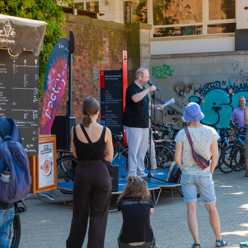 Mann tr&auml;gt drau&szlig;en auf einer mobilen B&uuml;hne was vor, Menschen drumherum h&ouml;ren zu. 