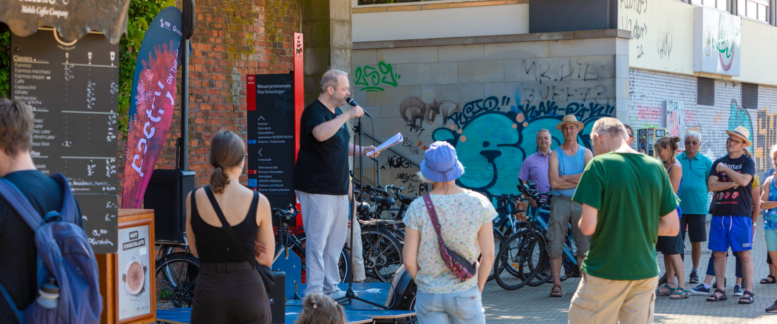 Poetry on the Road 2023 Mann trägt draußen auf einer mobilen Bühne was vor, Menschen drumherum hören zu.