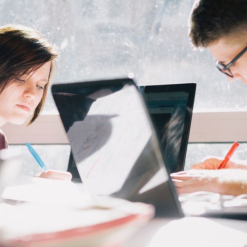 Zwei HSB Studierende sitzen an jeweils einem Laptop und schreiben handschriftlich auf daneben liegende Bl&ouml;cke. 