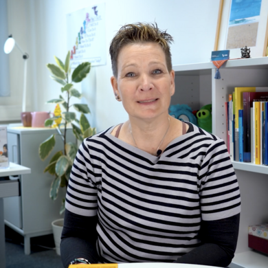 Auf dem Bild ist eine Frau mit kurzen braunen Haaren zu sehen. Sie hat ein gestreiftes Oberteil an und sitzt an einem Tisch in einem B&uuml;ro.