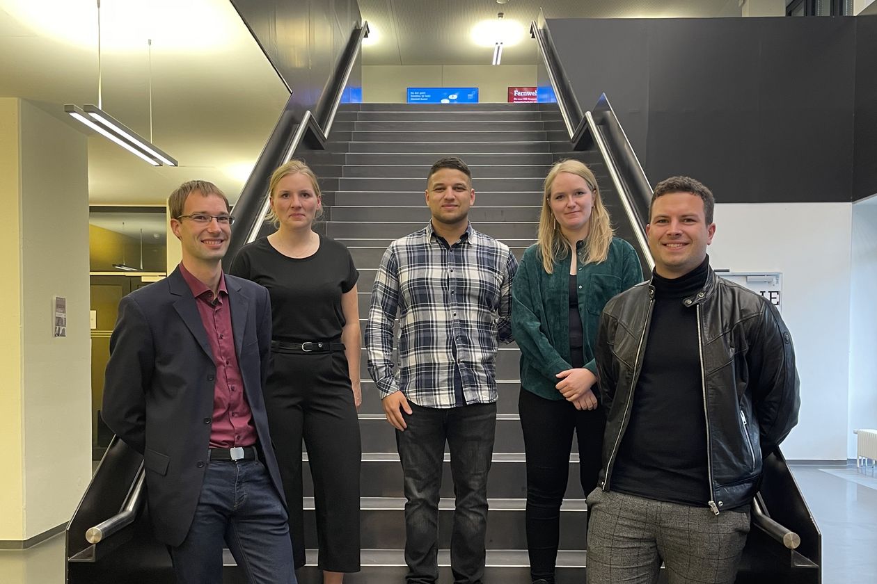 Fünf junge Menschen stehen an einer Treppe und lächeln in die Kamera.