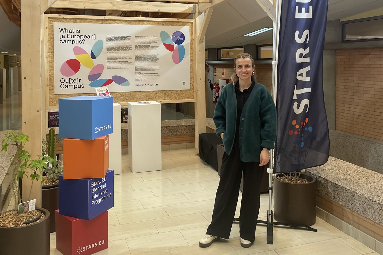 Janina Ebner STARS EU Projekt Outer Campus Junge Frau steht vor einem Pavillon aus Holz in einem Gebäude. Rechts neben ihr steht eine Fahne mit dem Schriftzug STARS EU. Lonks neben ihr stehen bunte Würfel.