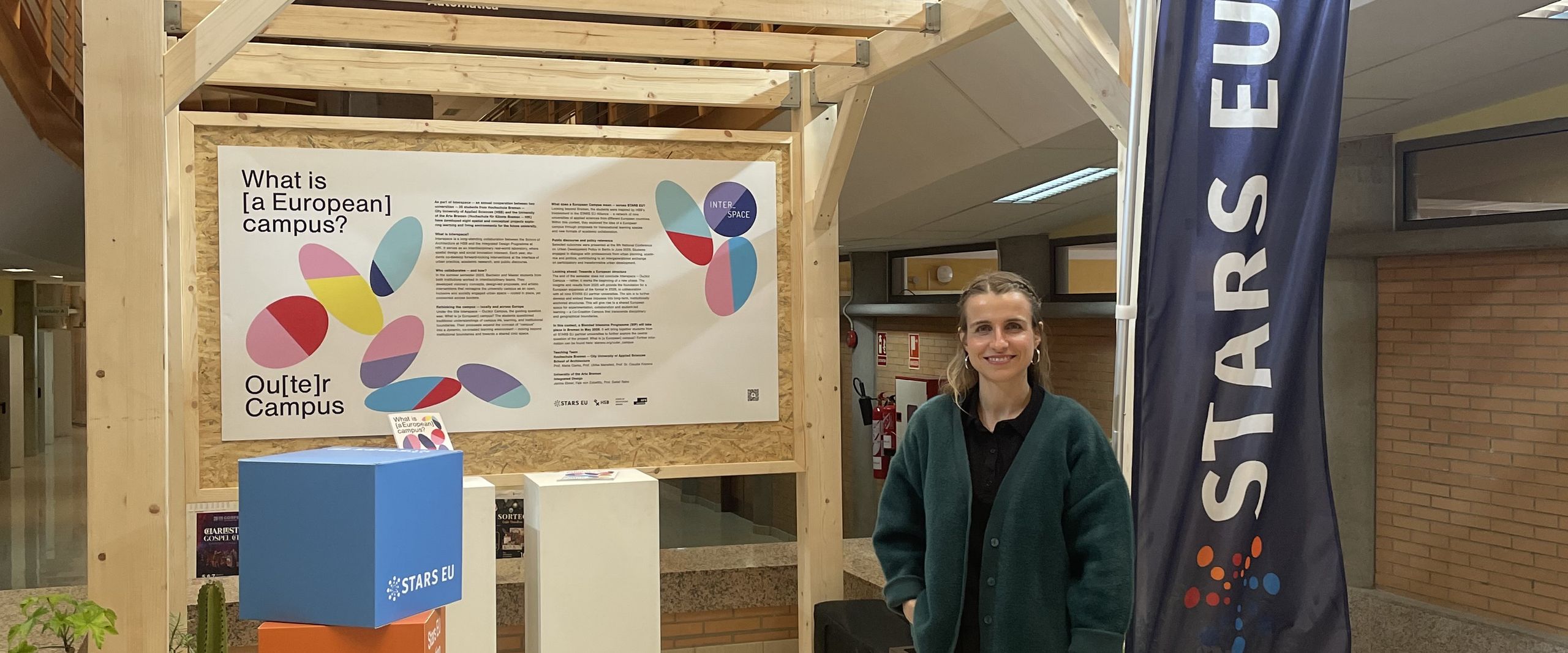 Janina Ebner STARS EU Projekt Outer Campus Junge Frau steht vor einem Pavillon aus Holz in einem Gebäude. Rechts neben ihr steht eine Fahne mit dem Schriftzug STARS EU. Lonks neben ihr stehen bunte Würfel.