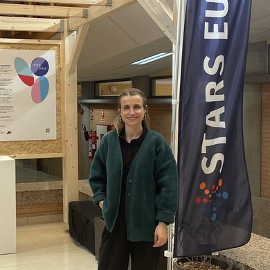 Janina Ebner STARS EU Projekt Outer Campus Junge Frau steht vor einem Pavillon aus Holz in einem Gebäude. Rechts neben ihr steht eine Fahne mit dem Schriftzug STARS EU. Lonks neben ihr stehen bunte Würfel.