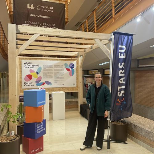 Junge Frau steht vor einem Pavillon aus Holz in einem Geb&auml;ude. Rechts neben ihr steht eine Fahne mit dem Schriftzug STARS EU. Lonks neben ihr stehen bunte W&uuml;rfel.
