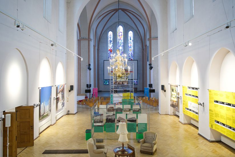 Das Bild zeigt den Innenraum einer ehemaligen Kirche, die als Ausstellungsraum genutzt wird, mit hohen Gew&ouml;lben, einem zentralen Kronleuchter, Stuhlreihen und gro&szlig;formatigen Informationstafeln an den W&auml;nden.