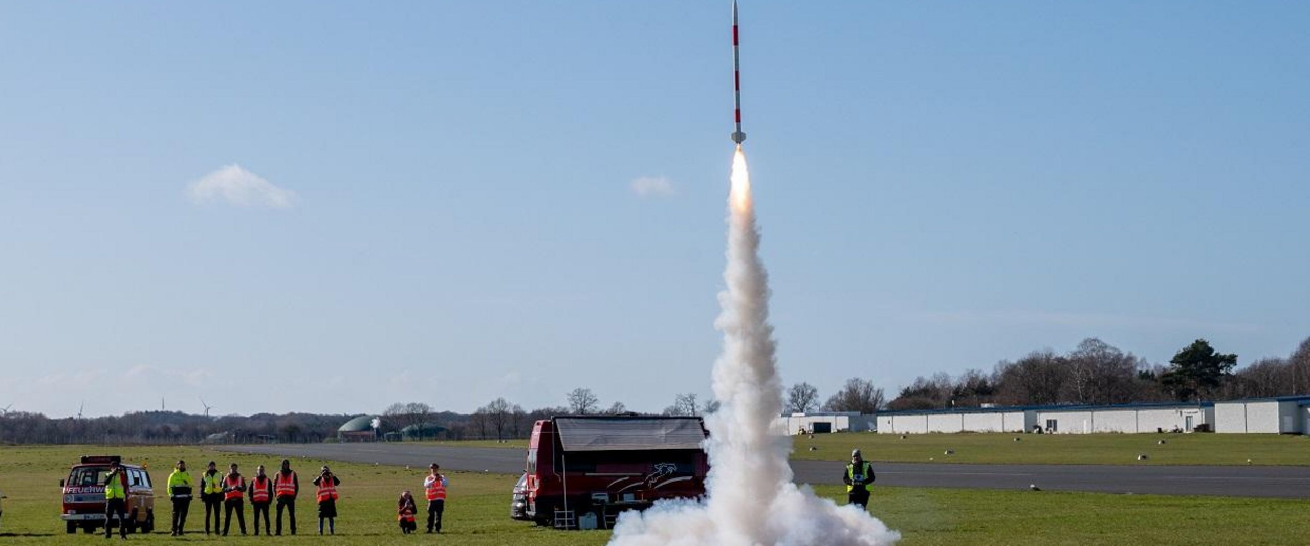 Auf einer Wiese startet eine Minirakete in den Himmel. Zuschauer:innen stehen dabei.