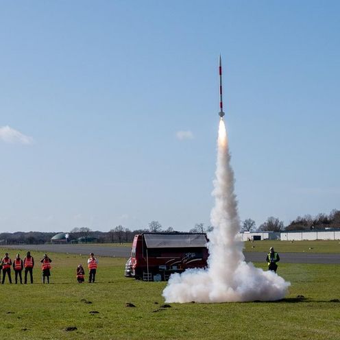 Auf einer Wiese startet eine Minirakete in den Himmel. Zuschauer:innen stehen dabei.
