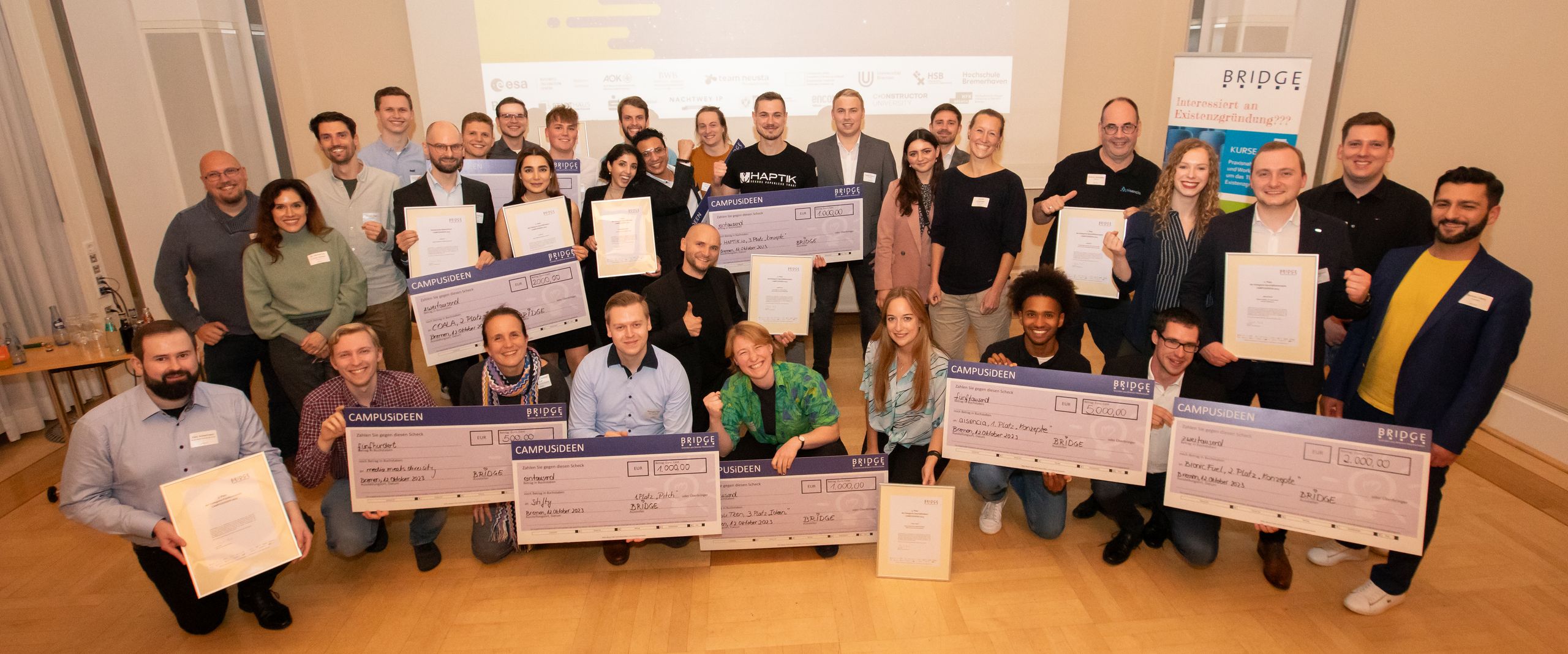 CAMPUSiDEEN 2023 BRIDGE Gruppenbild mit jungen Männern und Frauen. Sie halten jeweil übergroße Geldchecks