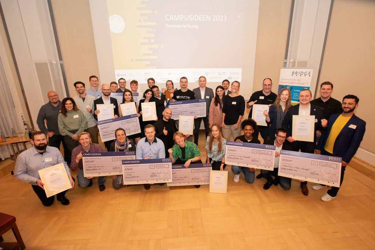 CAMPUSiDEEN 2023 BRIDGE Gruppenbild mit jungen Männern und Frauen. Sie halten jeweil übergroße Geldchecks