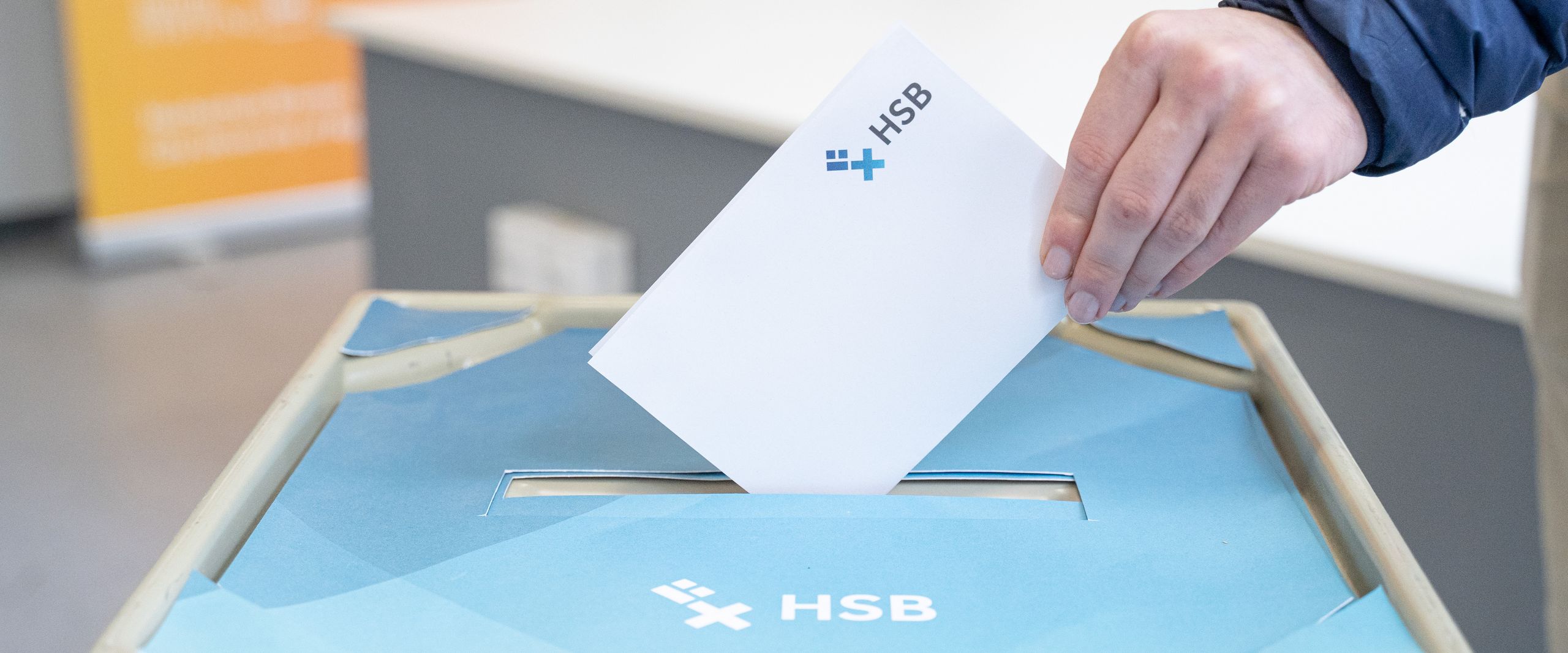 Wahlurne Auf dem Foto ist der Deckel einer Wahlurne im HSB-Design zu erkennen und eine Hand, die einen gefalteten weißen Zettel in die Wahlurne stecken will.