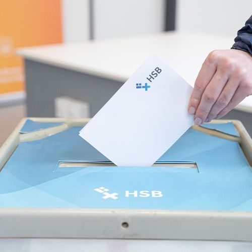 Auf dem Foto ist der Deckel einer Wahlurne im HSB-Design zu erkennen und eine Hand, die einen gefalteten wei&szlig;en Zettel in die Wahlurne stecken will. 