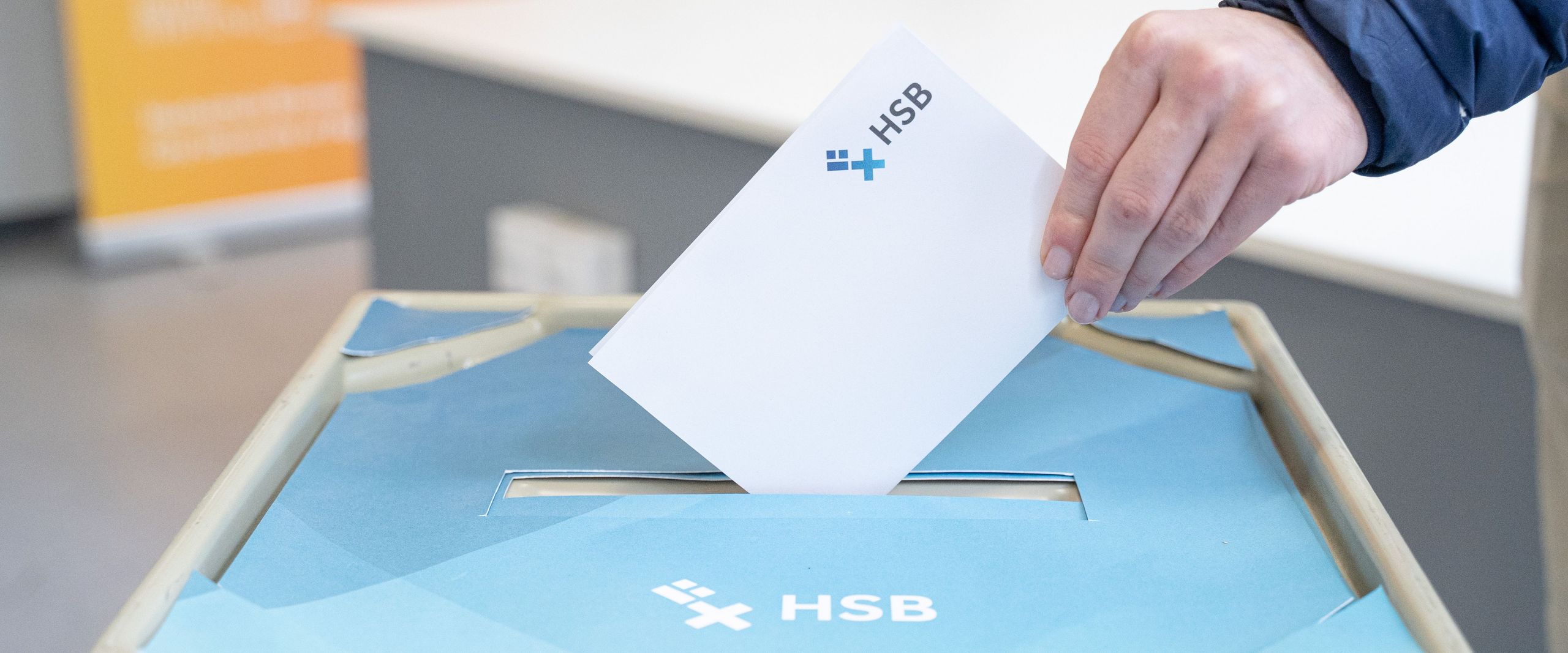 Auf dem Foto ist der Deckel einer Wahlurne im HSB-Design zu erkennen und eine Hand, die einen gefalteten weißen Zettel in die Wahlurne stecken will. 