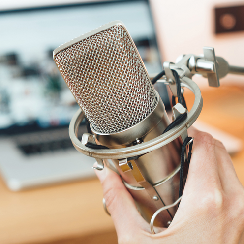Nahaufnahme von einem Mikrofon. Das Mikrofon wird von einer Hand gehalten. Der Hintergrund ist verschwommen. Im Hintergrund steht ein Laptop aufgeklappt auf einer Fl&auml;che. 