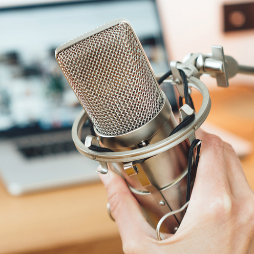 Nahaufnahme von einem Mikrofon. Das Mikrofon wird von einer Hand gehalten. Der Hintergrund ist verschwommen. Im Hintergrund steht ein Laptop aufgeklappt auf einer Fl&auml;che. 