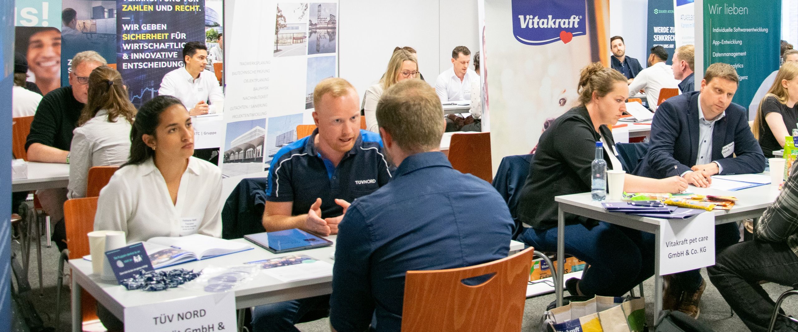 Männer und Frauen sitzen in einem Raum an Tischen und sind im Gespräch. Um sie herum stehen Roll-ups.