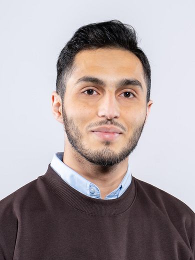 Auf dem Foto ist Rasheed Muhamad Ramadan zusehen. Er hat kurzes braunes Haar und einen kurzen Bart. Er tr&auml;gt ein hellblaues Hemd unter einem braunen Pullover. 