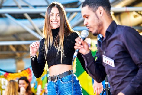 Eine Tänzerin und ein Sänger performen auf der Bühne. Hinter ihnen eine Brasilien-Flagge.