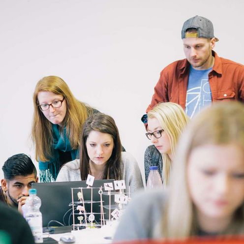 Auf dem Bild ist eine Gruppe Studierender zu sehen, die sich rund um einen Laptop positionieren und nachdenklich aussehen. Im Vordergrund sitzt eine Studentin vor einem roten Laptop. Der Vordergrund ist unscharf. 