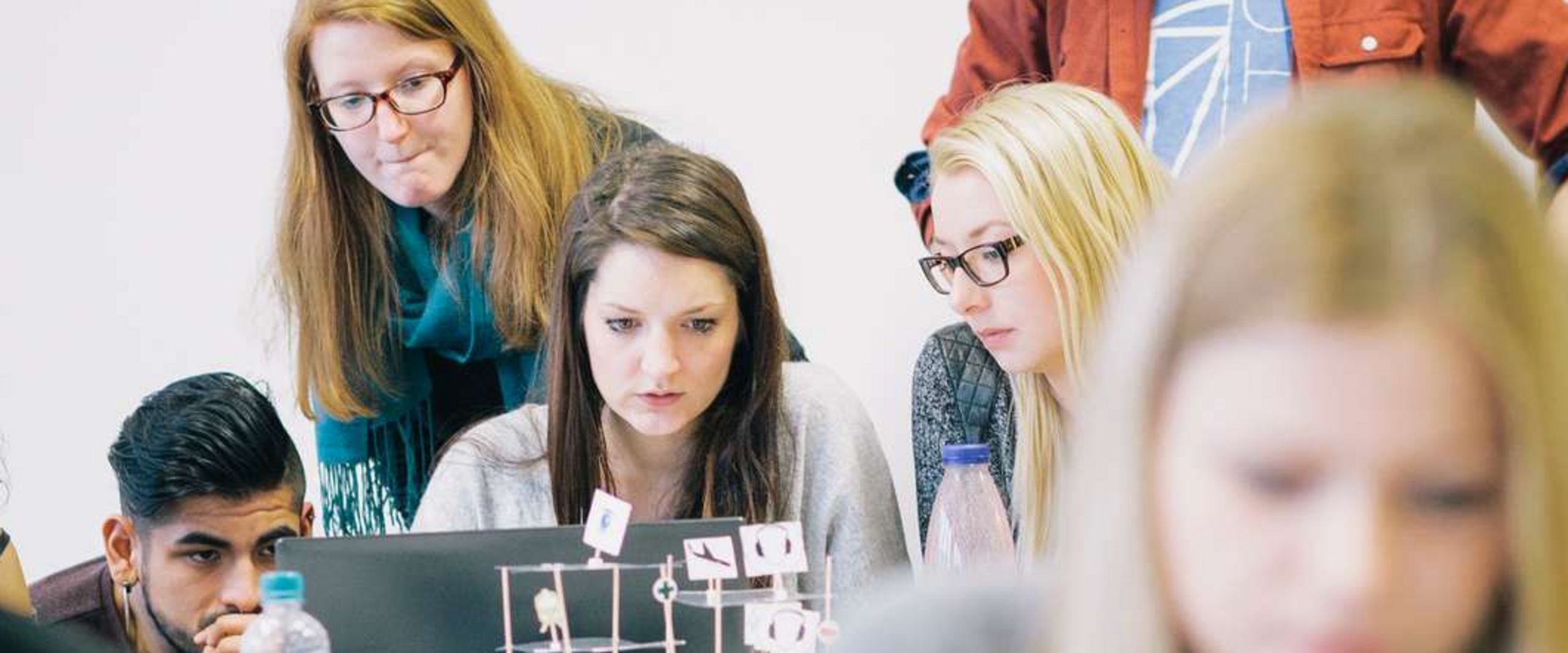 Gruppenarbeit von Studierenden Auf dem Bild ist eine Gruppe Studierender zu sehen, die sich rund um einen Laptop positionieren und nachdenklich aussehen. Im Vordergrund sitzt eine Studentin vor einem roten Laptop. Der Vordergrund ist unscharf.