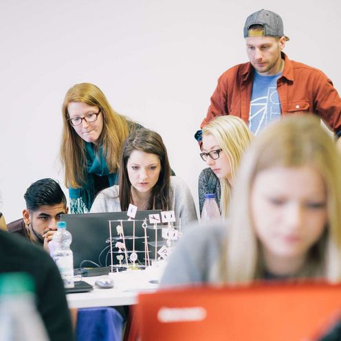Auf dem Bild ist eine Gruppe Studierender zu sehen, die sich rund um einen Laptop positionieren und nachdenklich aussehen. Im Vordergrund sitzt eine Studentin vor einem roten Laptop. Der Vordergrund ist unscharf. 