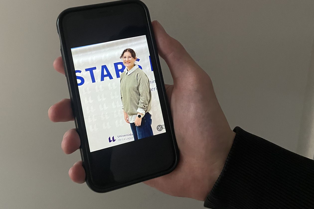 Hand holds mobile phone. On the screen one can see a young woman in front of a banner.
