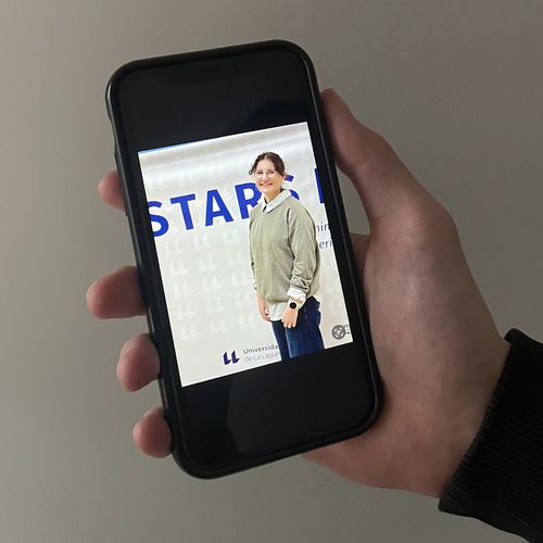 Hand holds mobile phone. On the screen one can see a young woman in front of a banner.