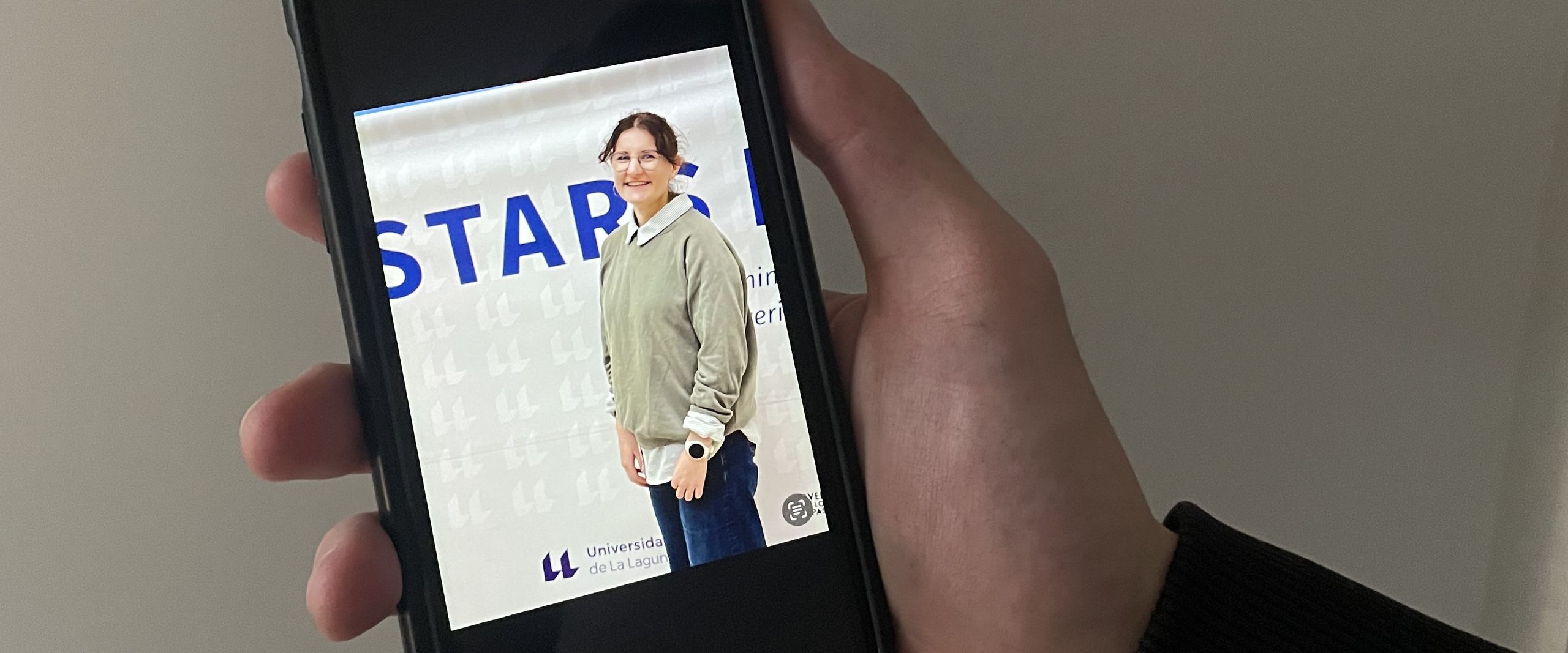 Hand holds mobile phone. On the screen one can see a young woman in front of a banner.