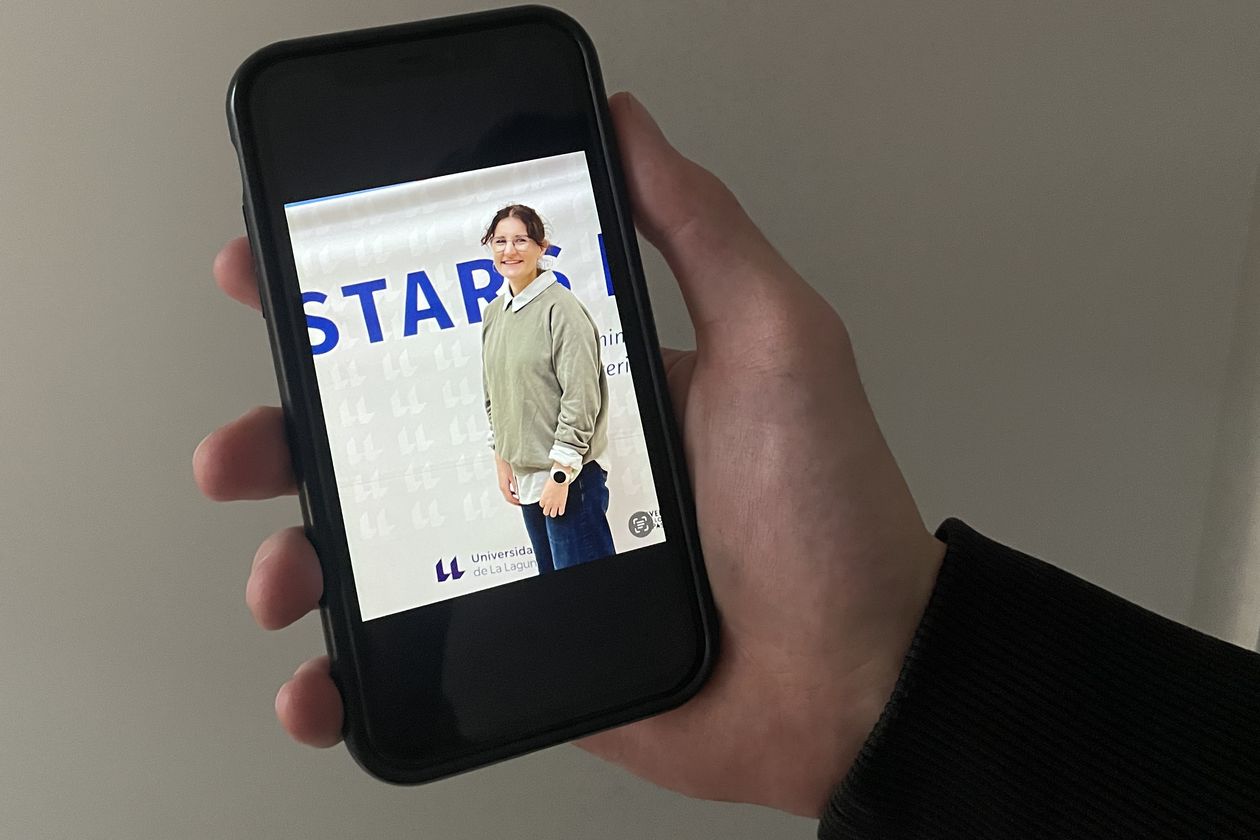 Hand h&auml;lt Handy. Darauf sieht man ein Foto einer jungen Frau vor einem Banner