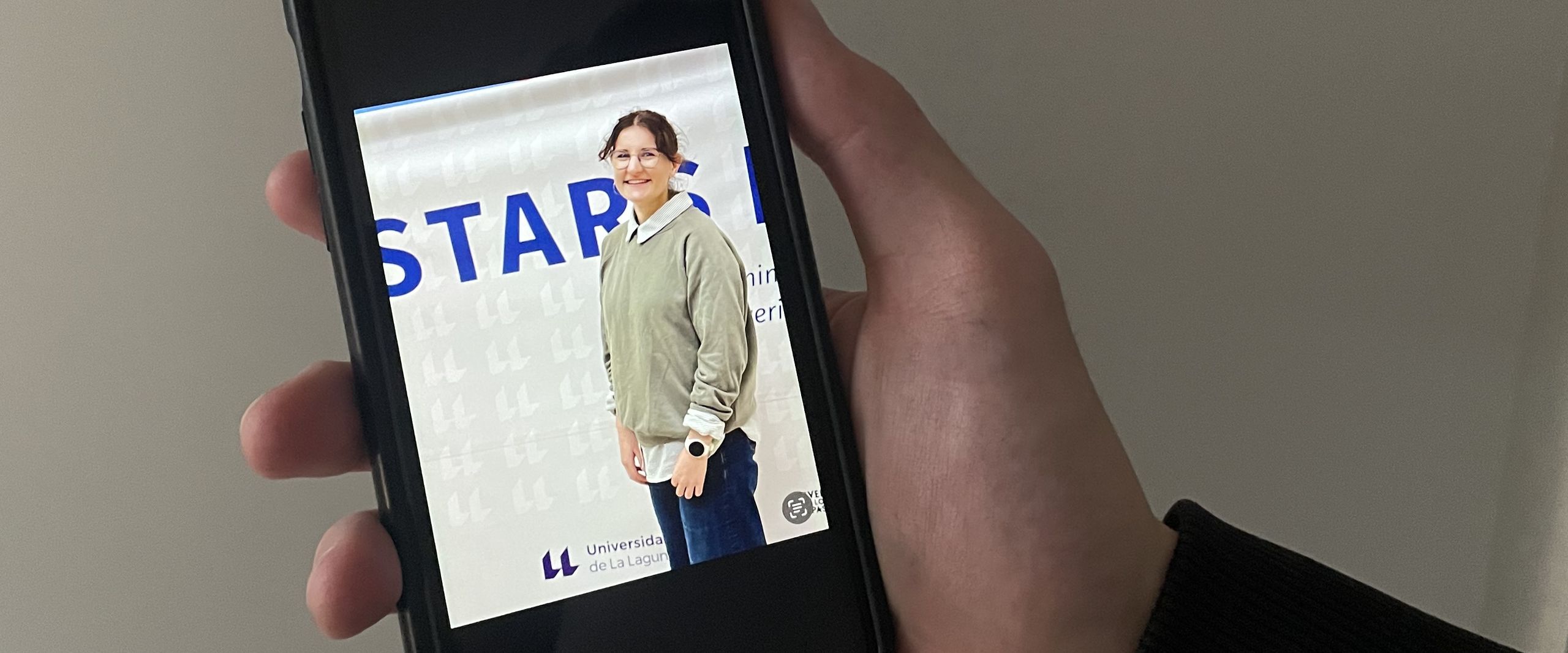 Hand h&auml;lt Handy. Darauf sieht man ein Foto einer jungen Frau vor einem Banner