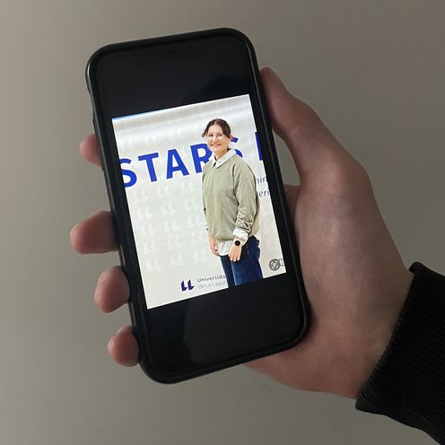 Hand h&auml;lt Handy. Darauf sieht man ein Foto einer jungen Frau vor einem Banner