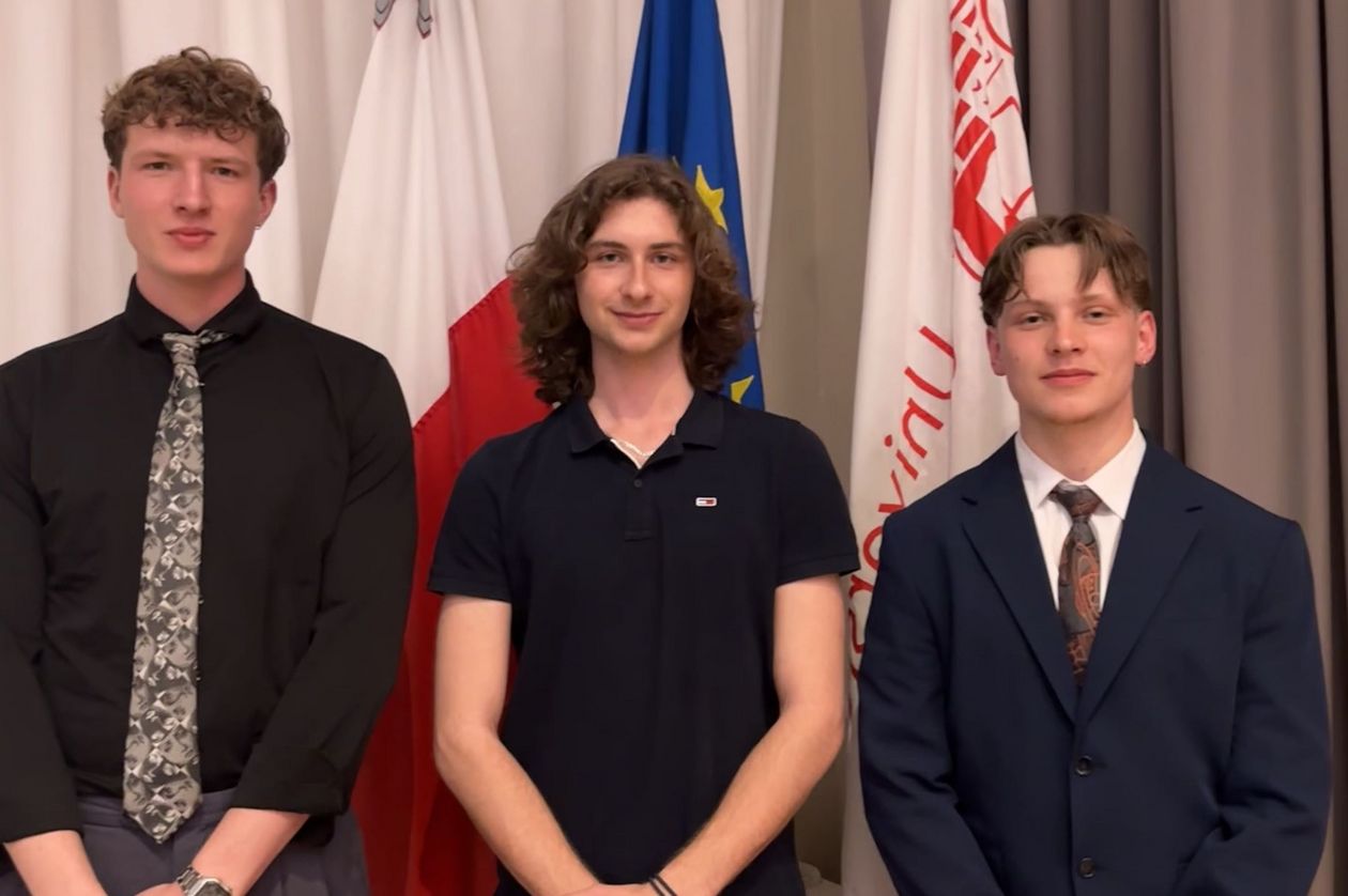 A group photo of three young men. They are standing in front of flags.