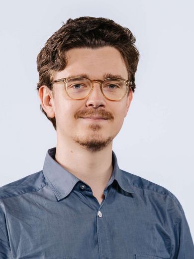Auf dem Bild ist Dirk Schmidt zusehen. Er hat braunes Haar und trägt eine runde Brille, einen Schnäuzer und ein blaues Hemd. 