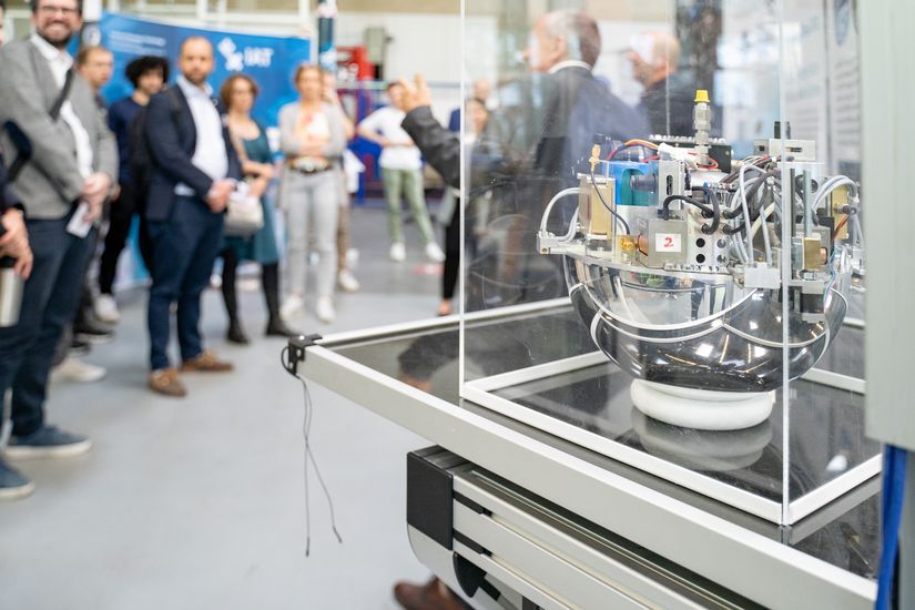 Auf dem Bild ist ein Modell eines Satelliten zusehen. Im Hintergrund ist unscharf eine Gruppe Personen zu sehen. Sie stehen im Halbkreis. 