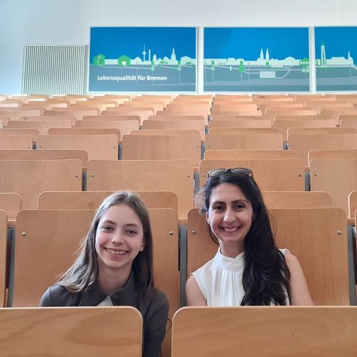 Mädchen und junge Frau sitzen nebeneinander in einem Hörsaal und lachen in die Kamera