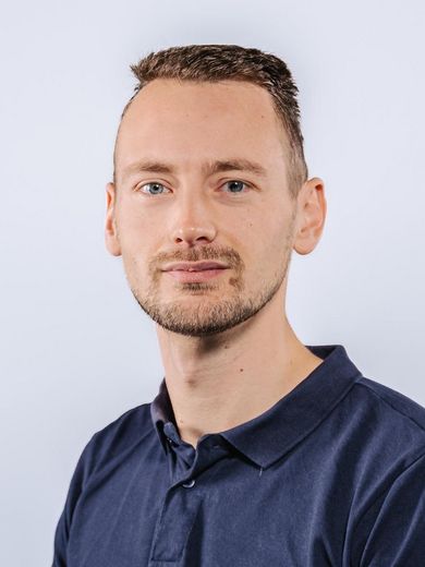 Auf dem Bild ist Alexander Grelle zusehen. Er hat kurzes braunes Haar und einen leichten Bart. Er trägt ein dunkelblaues Polo-Shirt. 