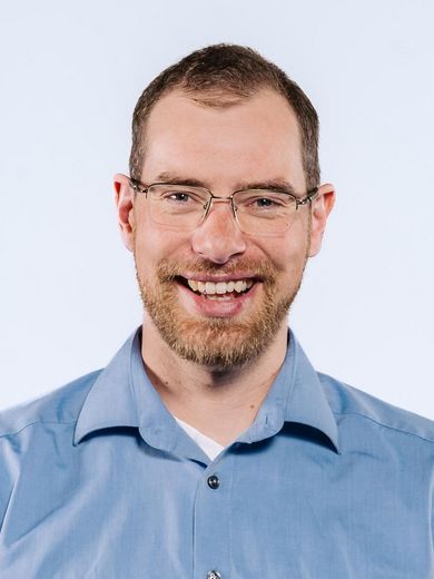 Auf dem Bild ist Philipp Tovar zusehen. Er hat kurzes braunes Haar und einen Bart. Er trägt eine unauffällige Brille und ein blaues Hemd. 