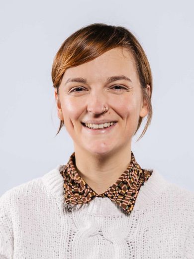 Auf dem Bild ist Lisa Kleiber zusehen. Sie hat ihr rotes Haar locker zusammengebunden. Sie trägt eine braungemusterte Bluse unter einem weißen Strickpullover. 