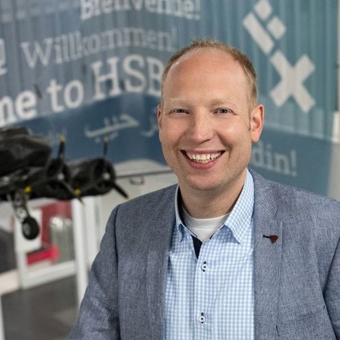 Auf dem Bild ist Jorgen von der Brelie zu sehen. Er tr&auml;gt ein blaues Sakko und ein hellblaues Hemd. Im Hintergrund ist ein Modellflugzeug in schwarz zusehen sowie ein Willkommensbanner der HSB in blau. 