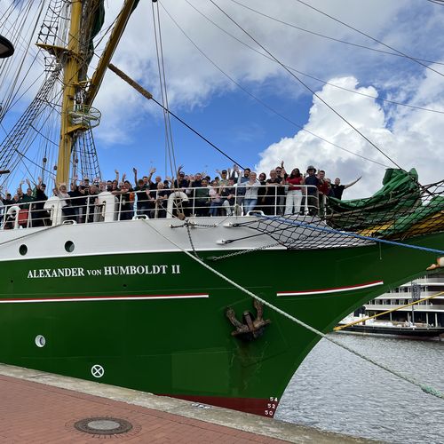 Segelschiff it gr&uuml;nem Rumpf. An Bord an der Spitze stehen Menschen und winken