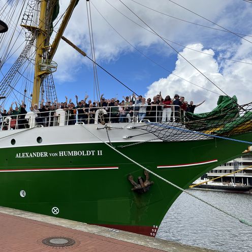 Segelschiff it gr&uuml;nem Rumpf. An Bord an der Spitze stehen Menschen und winken
