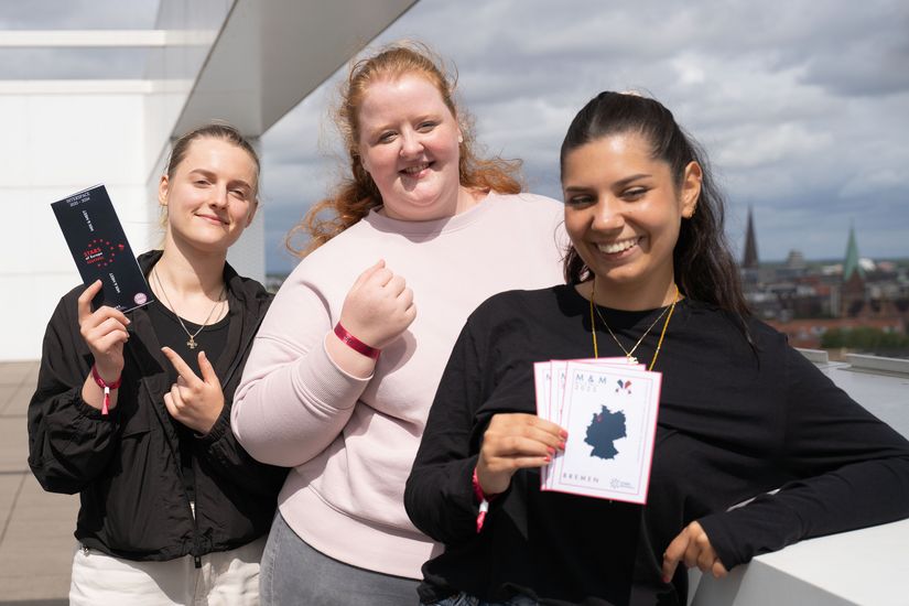 Three young women laugh into the camera. They are holding flyers.