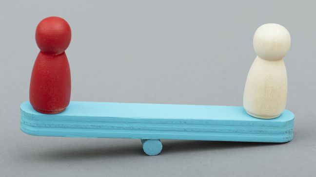 Ungleichgewicht Zwei Spielfiguren steehn auf einer Wippe. Die eine Seite ist unten, die andere oben.