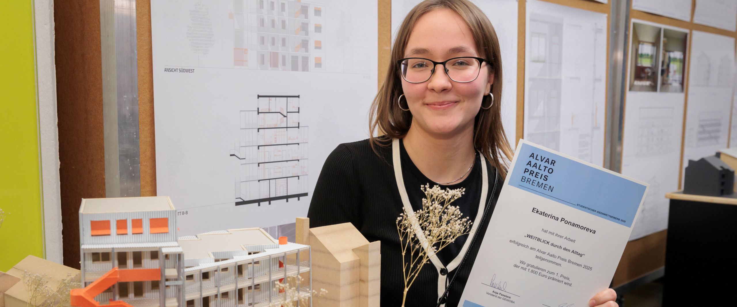 Junge Frau hält Urkunde und steht vor einem kleinen Holzmodell