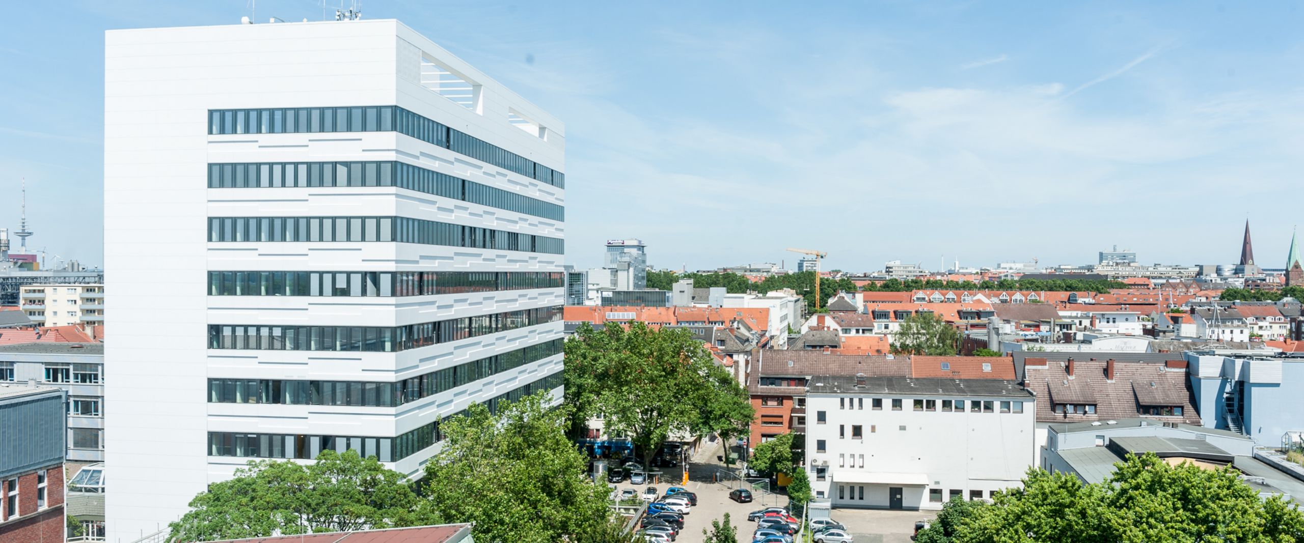 Luftaufnahme Campus Neustadtswall Auf dem Bild ist das AB-Gebäude des Campus Neustadtswall zusehen. Das Foto ist aus der Luft aufgenommen, sodass man außerdem noch blauen Himmel, grüne Bäume und einige Nebengebäude und den Parkplatz erkennen kann.