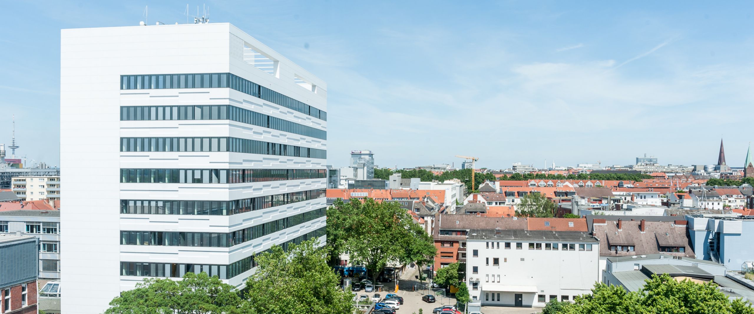 Auf dem Bild ist das AB-Gebäude des Campus Neustadtswall zusehen. Das Foto ist aus der Luft aufgenommen, sodass man außerdem noch blauen Himmel, grüne Bäume und einige Nebengebäude und den Parkplatz erkennen kann. 