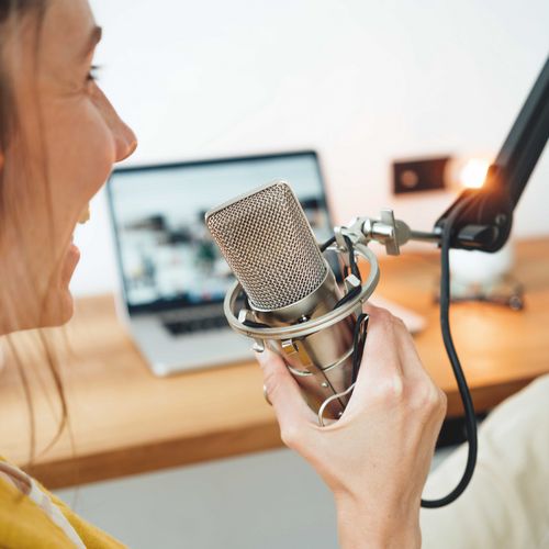 Foto einer Person, die in ein Mikrofon spricht, im Hintergrund ein Laptop