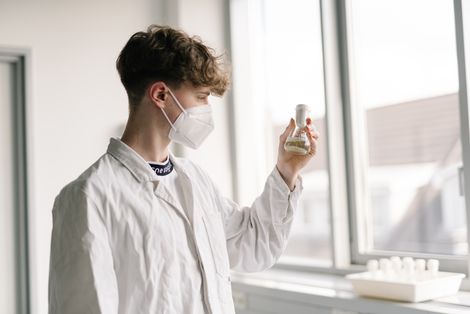 Student mit Maske steht im Labor und schaut sich ein chemisches Mittel an.